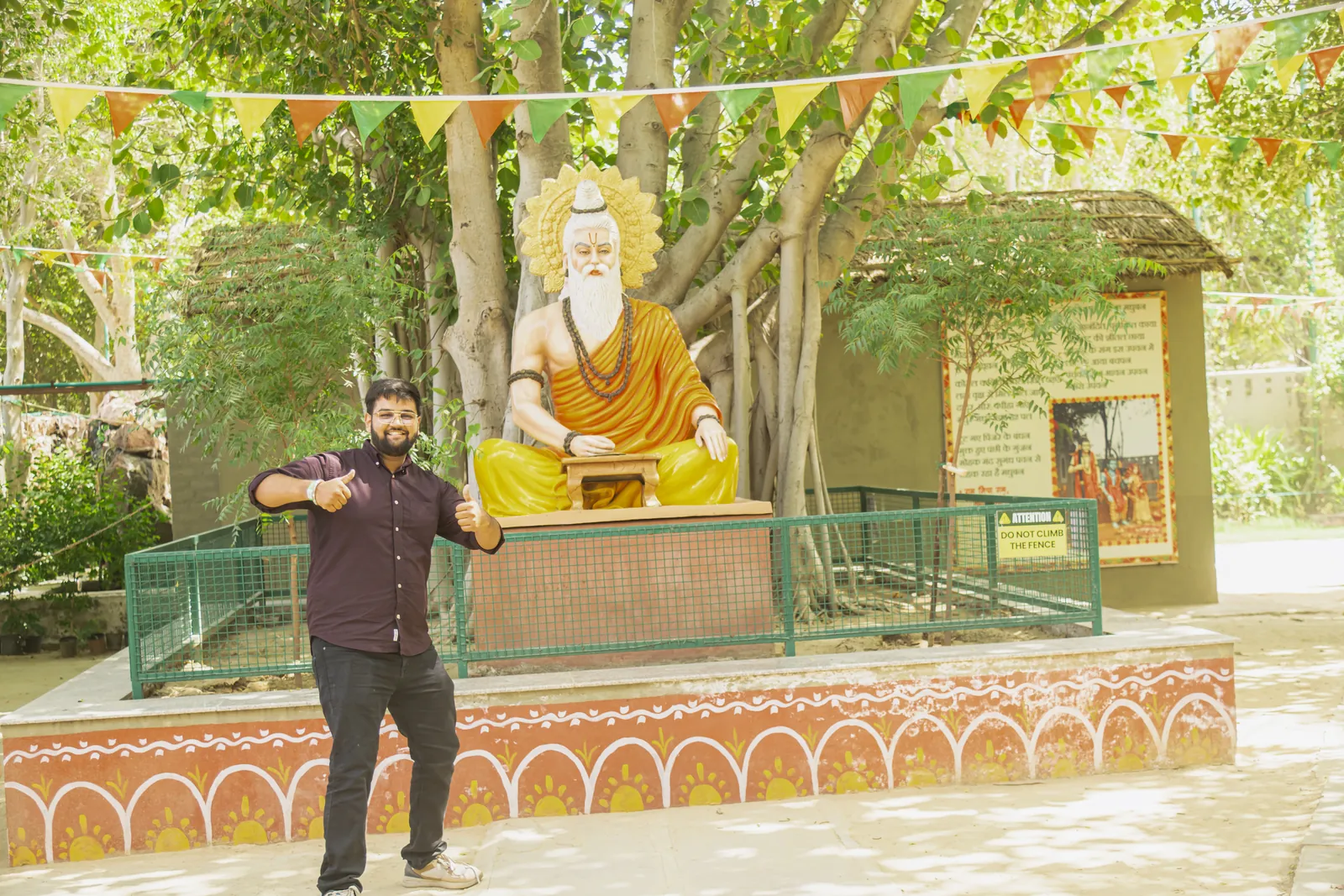 A large painted statue of a seated white-bearded sage in saffron robes on a decorated plinth among the trees, with strings of flags overhead and a visitor standing in front of it