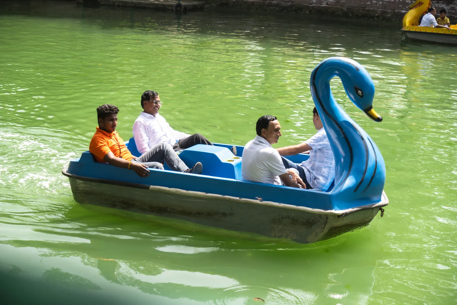 Four guests pedalling a blue swan-shaped pedal boat across the green water of the lake at Pratapgarh Farms, with two more boats behind them