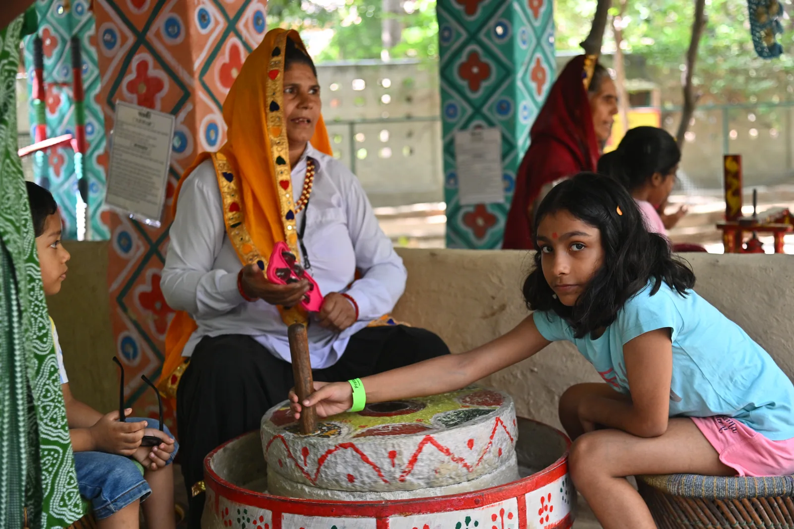 A girl turning the stone chakki in the village yard while a woman in an orange odhni holds the grain and two children watch