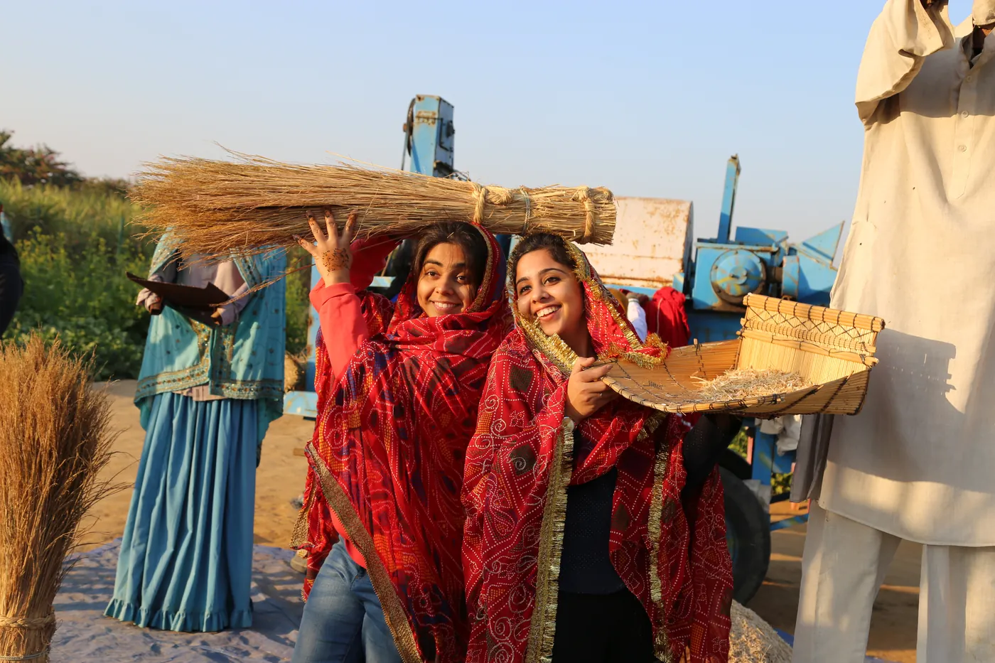 Two women in red printed dupattas holding a bundle of harvested stalks and a winnowing basket beside a threshing machine