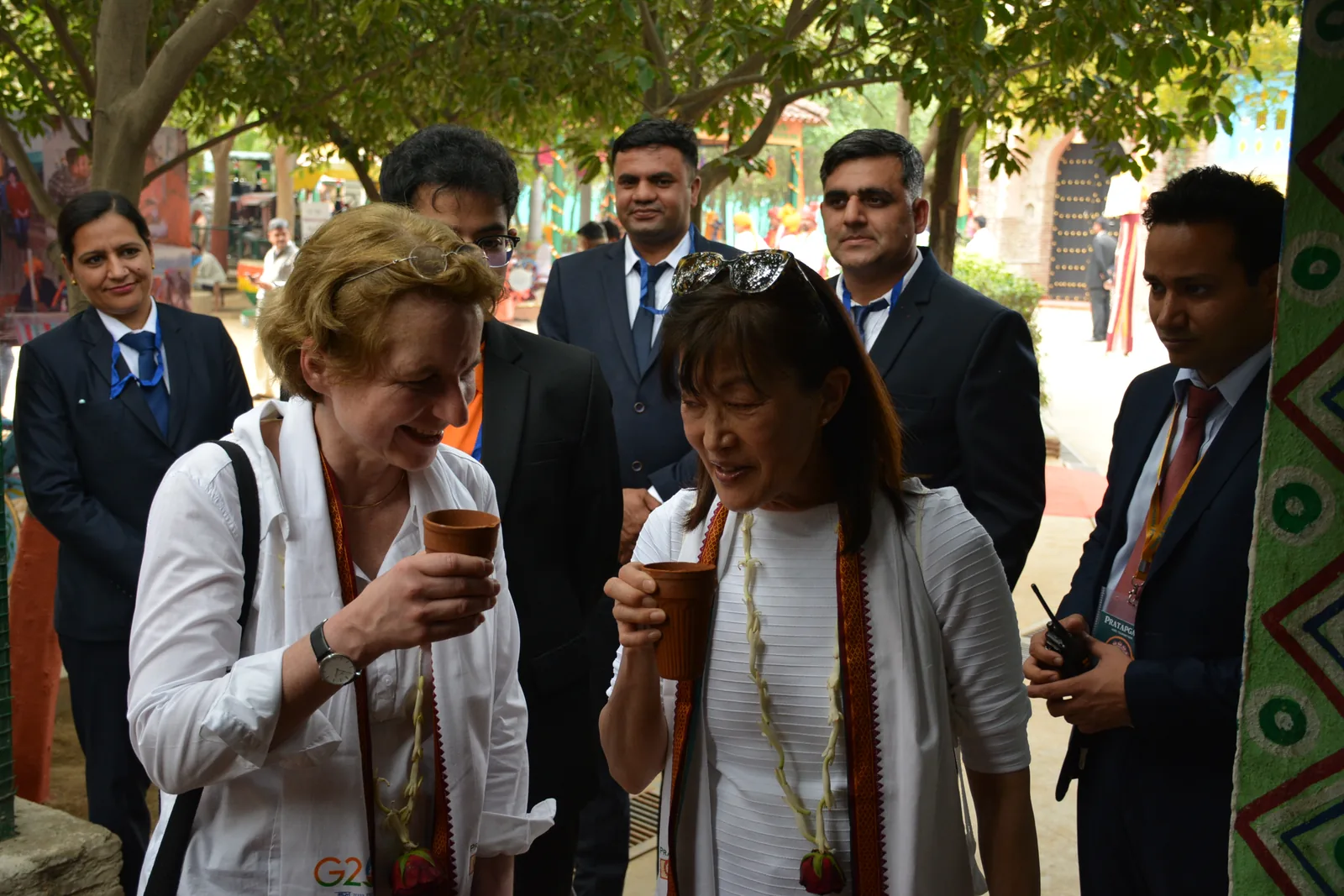 Two delegates drinking buttermilk from terracotta kulhads and smiling, with other delegates in suits behind them