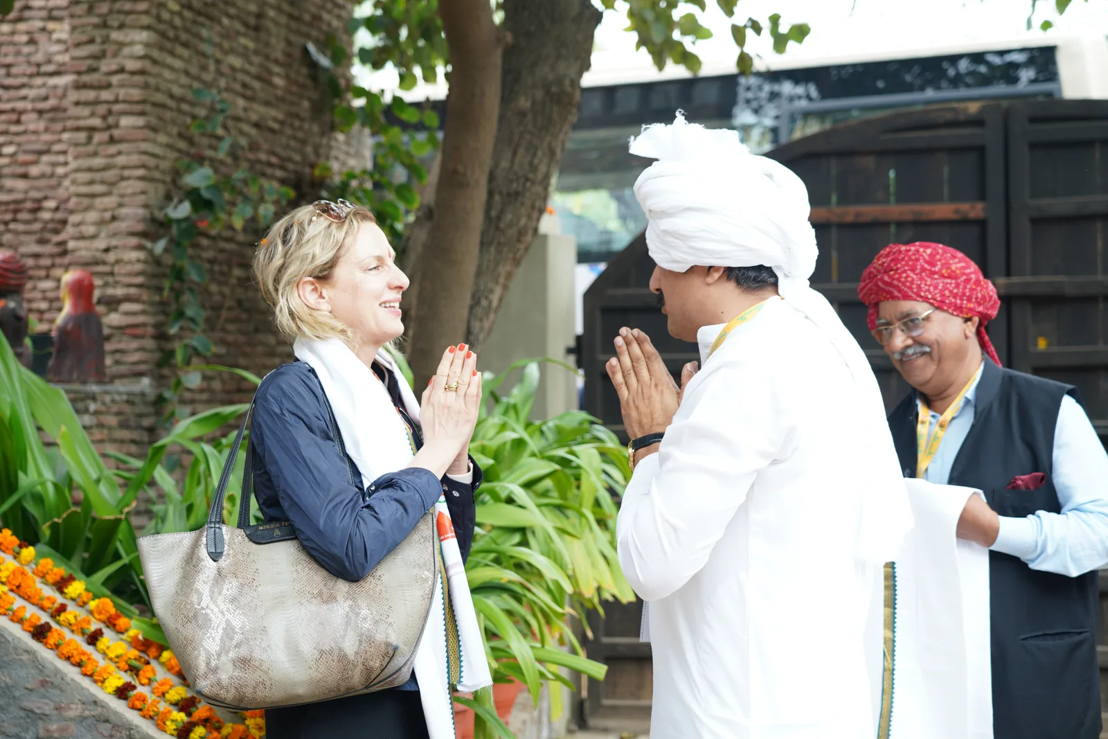 A delegate in a navy jacket with folded hands being welcomed at the farm gate by two men in Haryanvi pagdis, with marigolds along the wall