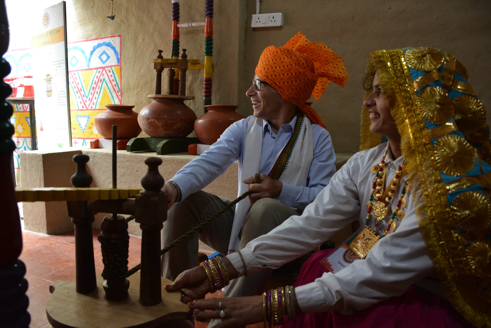 A delegate and a host seated together at a wooden bilona churn, both pulling the rope, with terracotta pots stacked on the wall behind